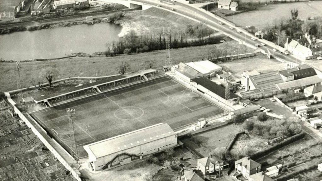 A black and white aerial photo of Cambridge United's Abbey Stadium between the 1930s to possibly 1950 showing the ground with two stands, four tall light towers in each corner, allotments to its left and above it a park and Newmarket Road 