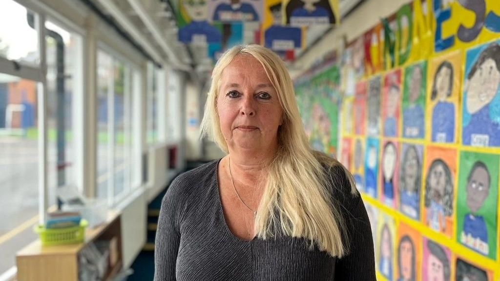 Helen Slack is front and centre looking at the camera in a school corridor with pictures on the walls. She has long blonde hair and is wearing a dark grey top