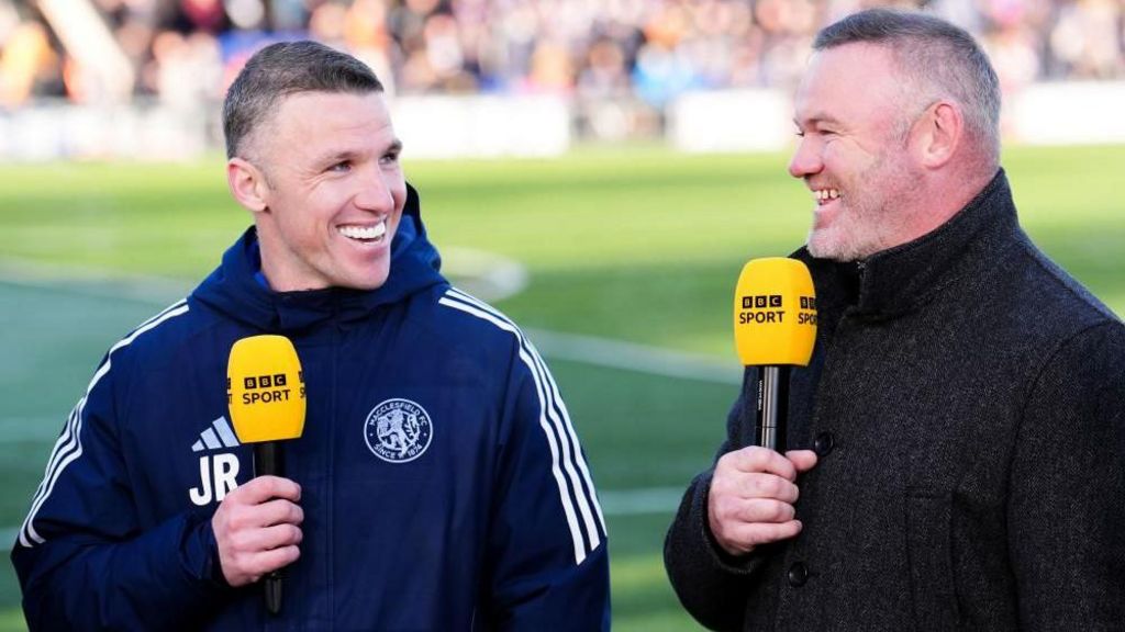Two men are smiling while standing next to each other by the football pitch. They are both holding BBC Sport microphones.