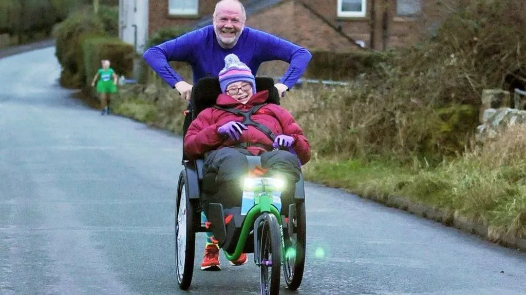 Chloe is sitting in a custom wheelchair wearing a pink coat and a purple hat and is being pushed by her dad Stephan who is wearing a purple long-sleeved top