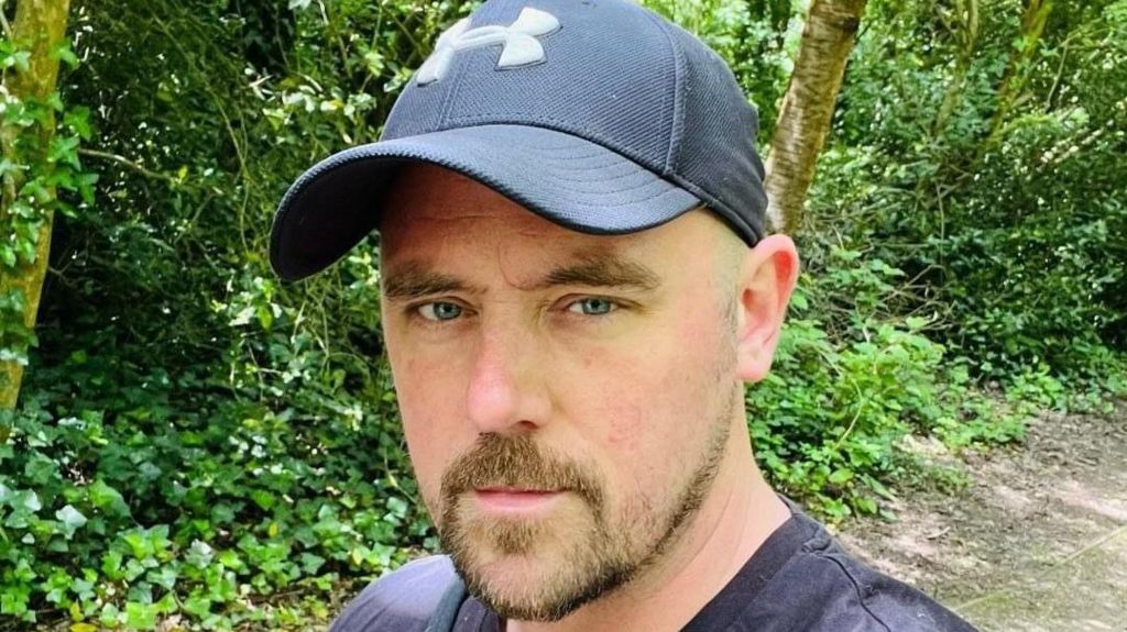 Andy Wilson looks at the camera, wearing a blue baseball cap and dark T-shirt, with trees and shrubs and a path behind him