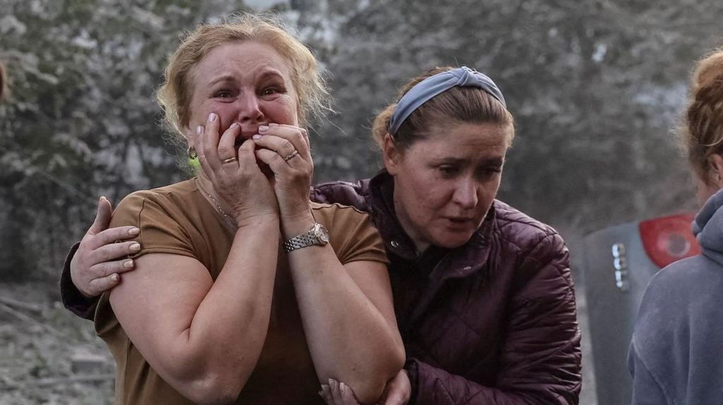 A woman in a burgundy coat holds another woman in a brown t-shirt near an apartment building hit by a Russian missile strike in Lviv. She holds her hands to her face in distress.