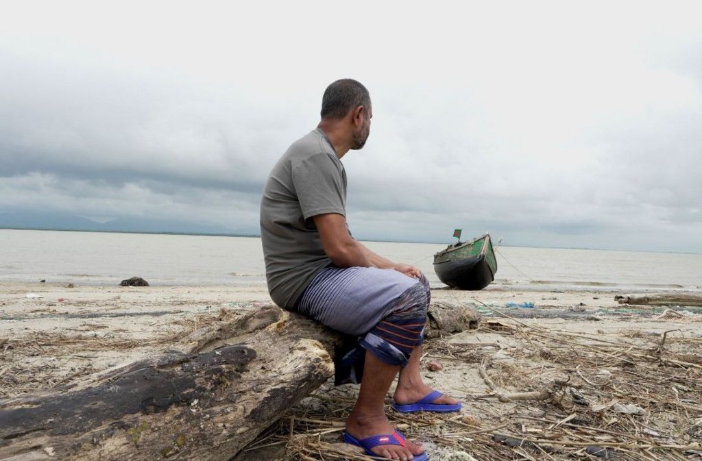 Nasir sits on a log on the bank of a river looking out across the river. His face is turned away and is not visible. He is wearing a grey t-shirt, shorts, and sandals. The day is overcast, and a small boat is also visible on the bank of the river. 