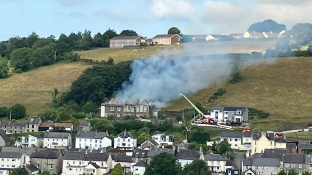 A number of houses with hilld and the school building in the background. There is a lot if smoke coming from the building and fire vehicles parked at it