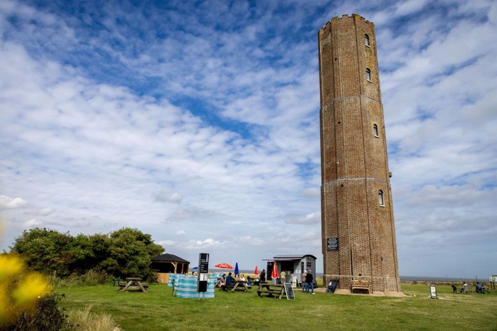 The Naze Tower in Walton-on-the-Naze. It is 86ft (26m) tall, made of brown bricks and stands in front of a cloudy blue sky. At its base are a number of benches and a food hut.