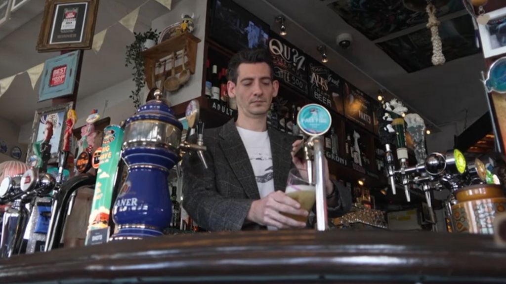 A dark-haired man pulls a pint with a bar in the background. The picture is taken from a low angle