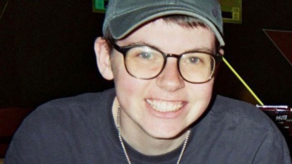 Alex, a 21-year-old man, smiling at the camera. He is wearing a grey baseball cap and a grey t-shirt. He has glasses.