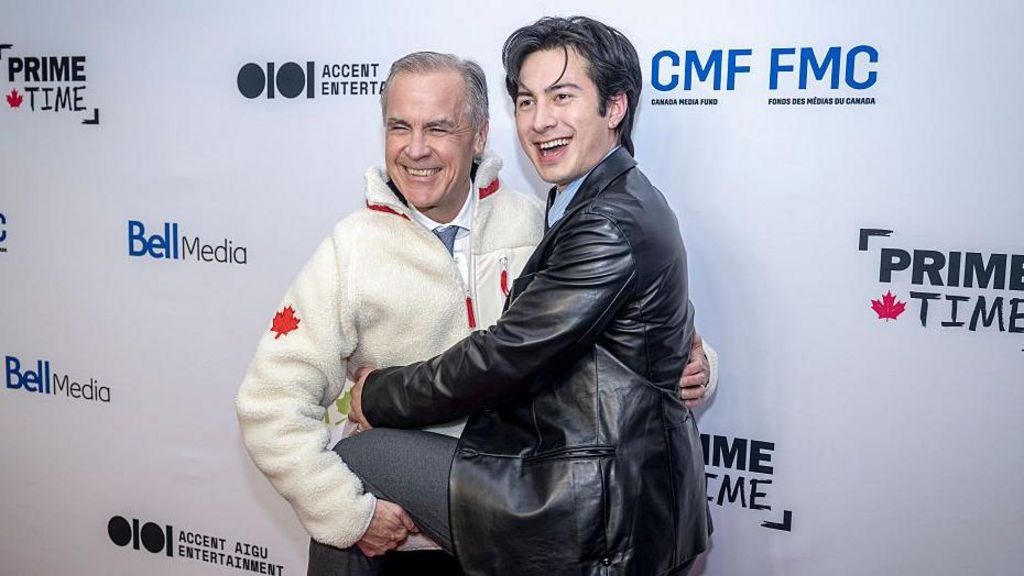 Prime Minister Mark Carney, wearing a beige fleece sweater with a red Canadian maple leaf on the arm, laughs as he hugs Heated Rivalry actor Hudson Williams, wearing a leather jacket.