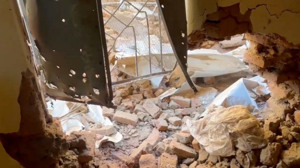 Bricks and rubble lie on the floor inside a room. The walls are cracked and the shutters that once covered the window are bent and riddled with holes.