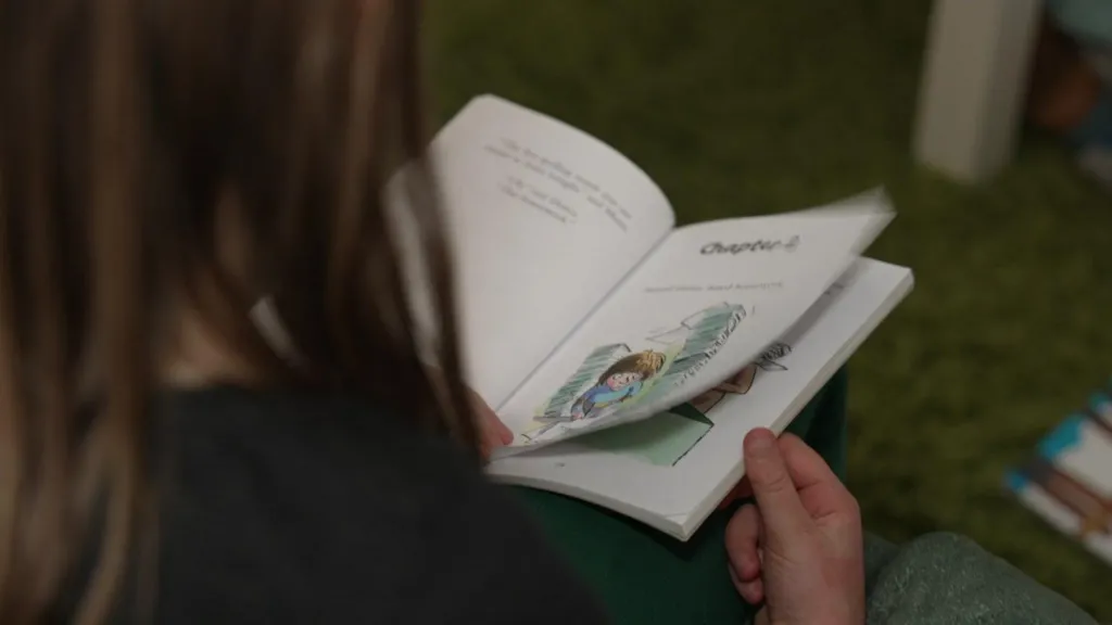 The picture shows a child sitting on a soft, green carpeted floor and reading a small illustrated book. The child is holding the book open on their lap, turning a page to a chapter that includes a colourful drawing of a character. Only the back of the child’s head, shoulders and hands are visible, so you cannot identify them. 