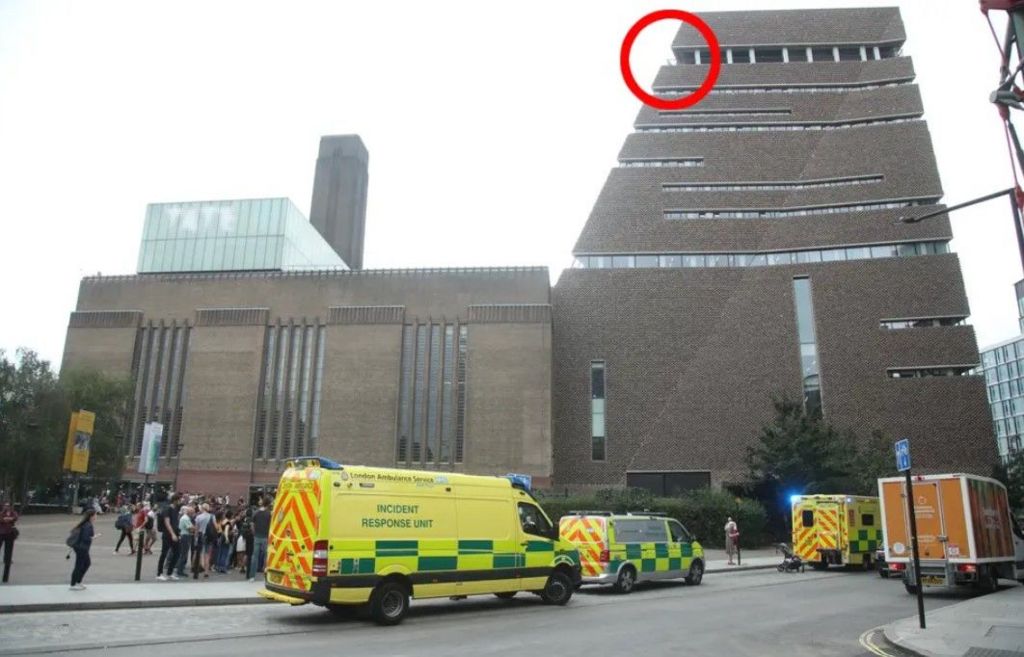 The Tate Modern building is pictured with ambulances outside. People are in the middle distance looking up. On the top floor of the brown-bricked building is a red circle to show where the boy fell from.