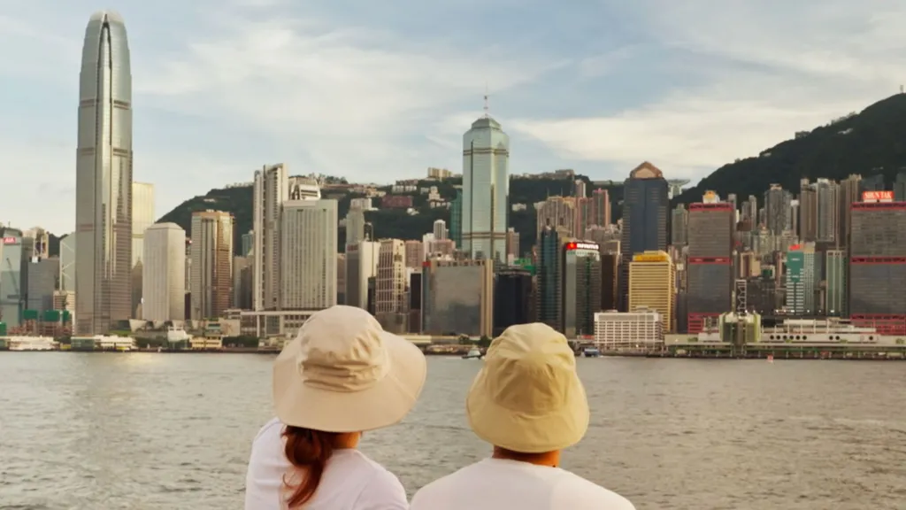 Eric and Emily shot from behind - both wearing light coloured bucket-style hats and white t-shirts. They are looking over the water at a city skyline.