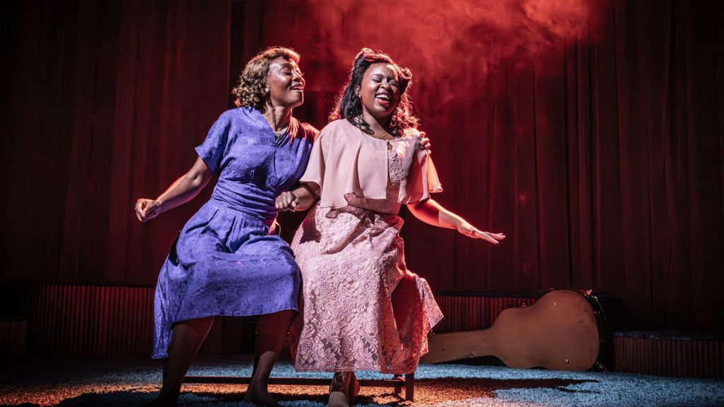 Beverley Knight in a 1940s style purple dress dancing with her co-star Ntombizodwa Ndlovu who is in a pink floral 1940s dress. They are on stage in Marie and Rosetta. 