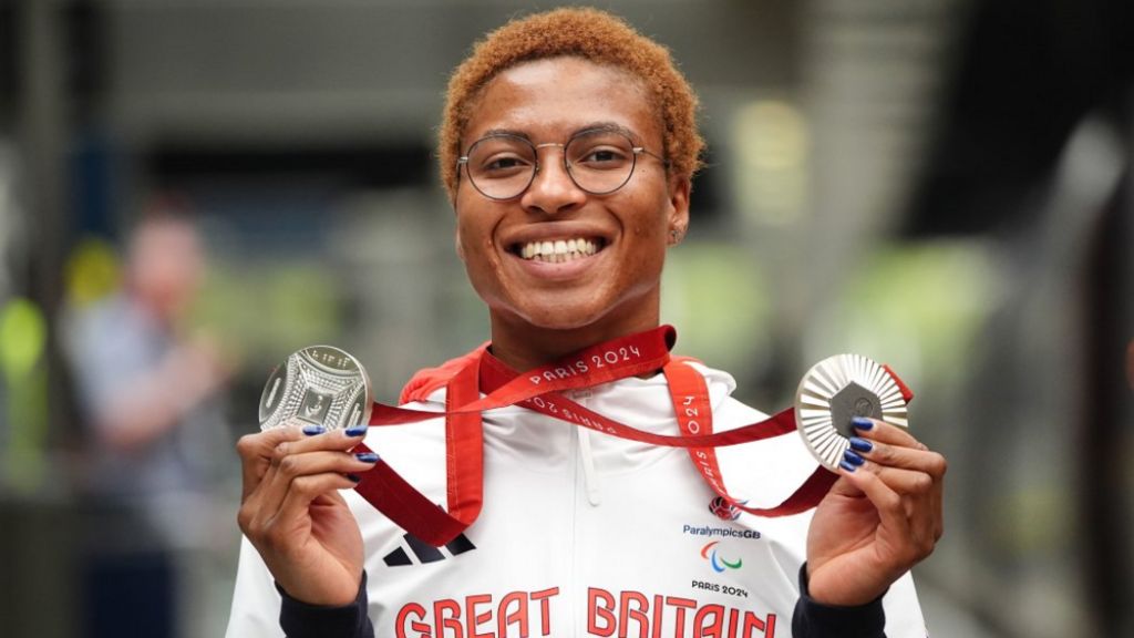 A woman in a Paralympics GB top smiles at the camera and holds up a silver medal in either hand