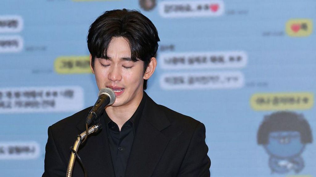 A tearful Kim Soo-hyun looking down and speaking into a microphone. He is wearing a black shirt and black blazer. Behind him is a projection of screenshots of text messages.