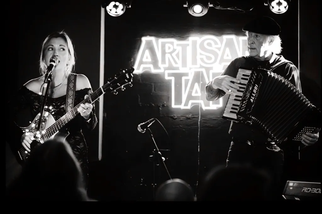 A female and male artist on stage at the Artisan Tap. She is playing a guitar and singing into a microphone. He is watching her and playing an accordian. 