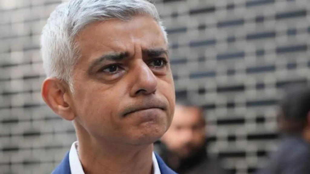 Sir Sadiq Khan, Mayor of London stood outside with a puzzled face staring into the distance. He is wearing a blue blazer and open collared shirt.