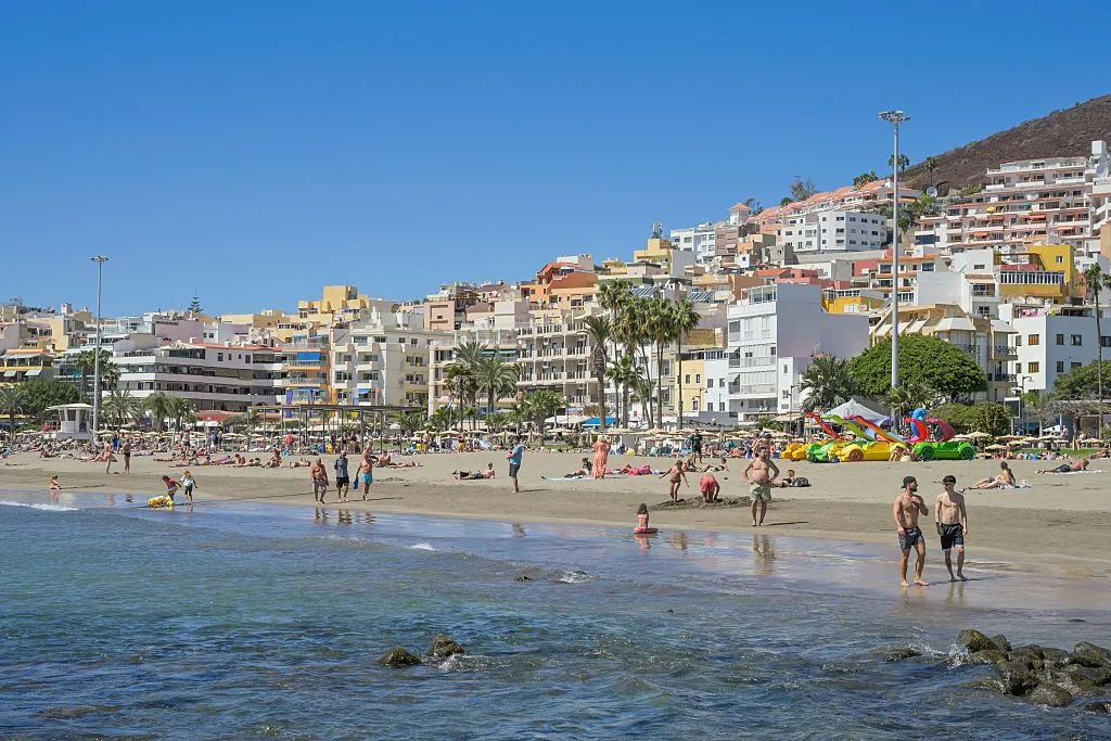 Locals and tourists swim, walk and play on a sandy beach. The water is clear and calm and there is a backdrop of palm trees and colourful apartment buildings in the background