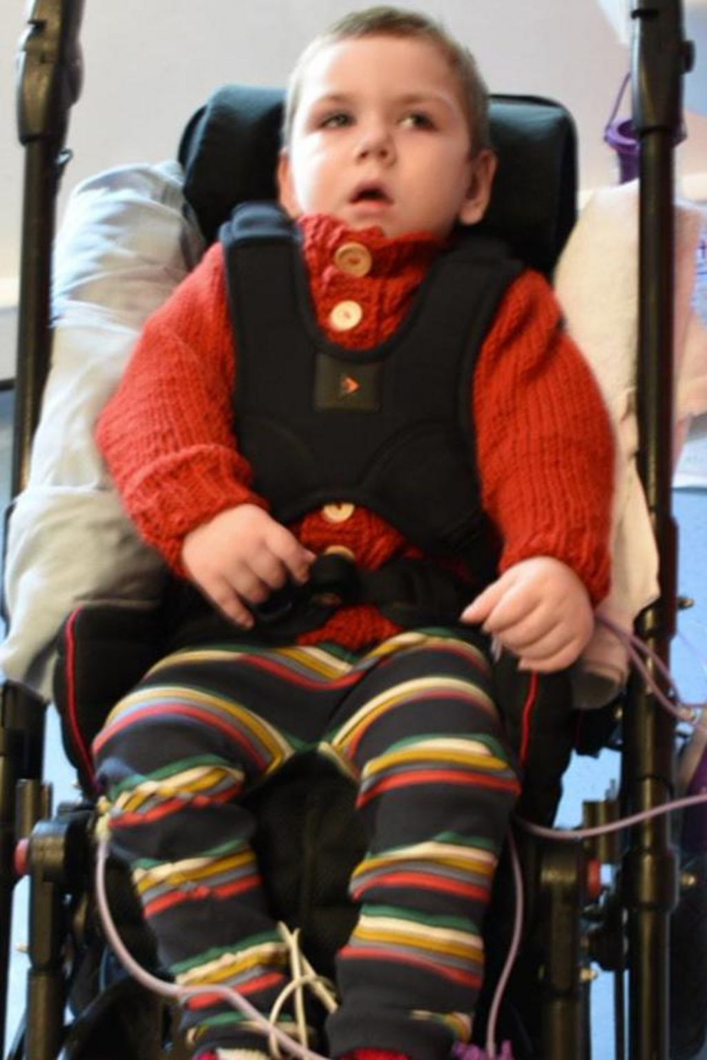 Kol Page appears to be sitting, strapped into either a child's wheelchair, or modified pushchair, with a pillow behind him. Medical tubes appear to be visible around his legs. He wears an orange jumper and striped trousers. 