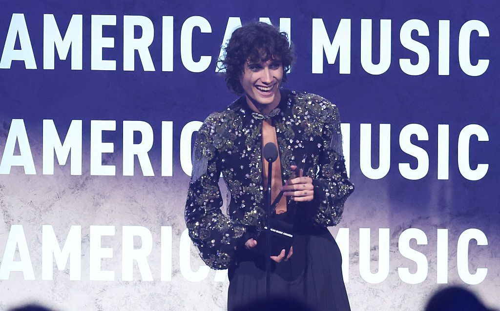Sombr collecting an award on stage - he is wearing black trousers and a dark glittery top slashed down the middle at the front.