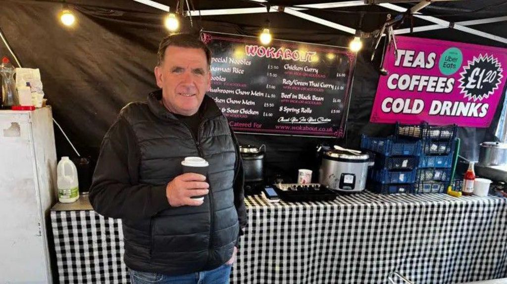 David is pictured standing in front of his "Wokabout" food stall, holding a black takeaway cup. The background shows a menu listing various Thai and Chinese dishes and prices.