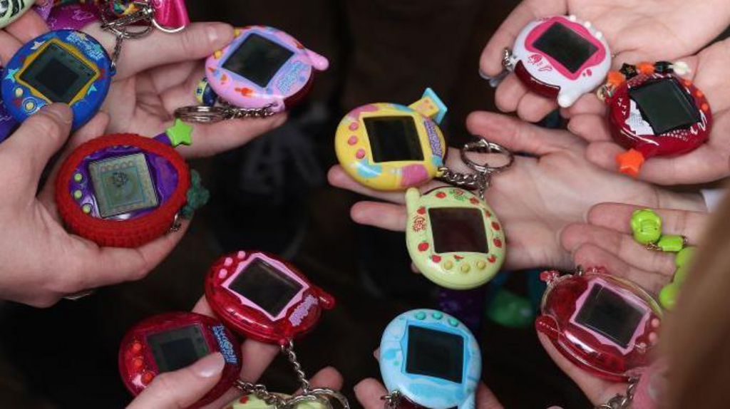 Tamagotchi in lots of different colours.  They are small egg-shaped devices with a digital screen in the middle and three buttons underneath. The colours range from a deep see-through red to bright blue, green, yellow and purple.