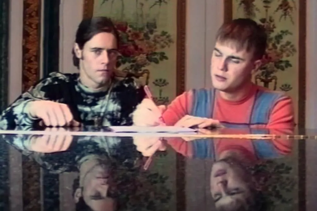 Howard Donald and Gary Barlow handwrite lyrics while sitting at a piano. Their faces are reflected in the piano lid.