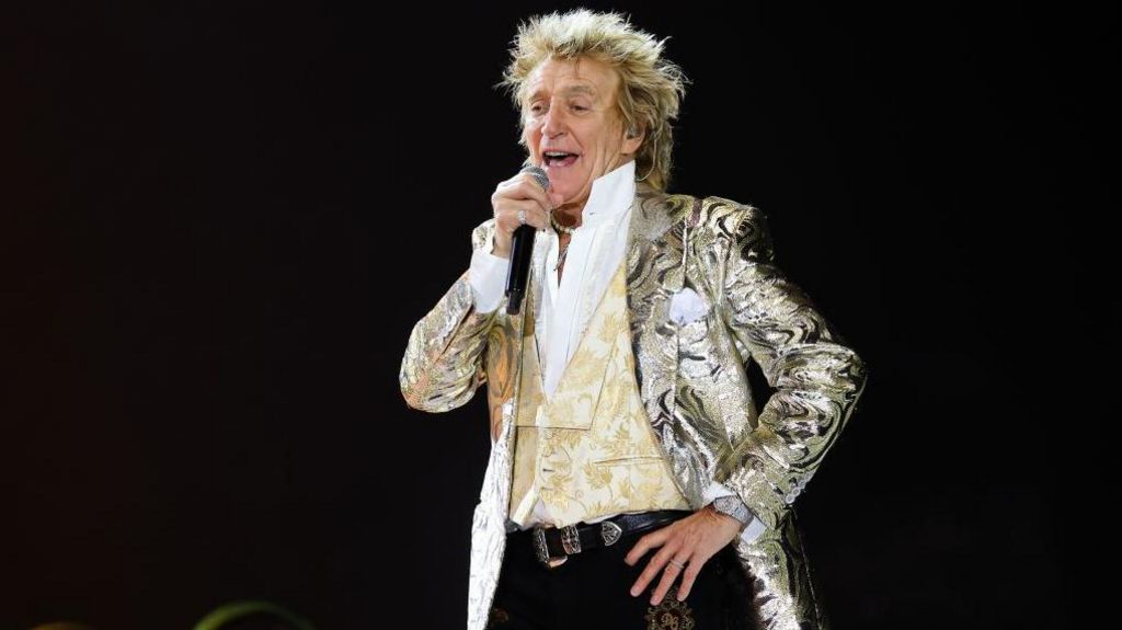 Rod Stewart talks into a microphone with one hand on his hip, while wearing a silver paisley coat and a brocaded waistcoat. He looks fabulous.