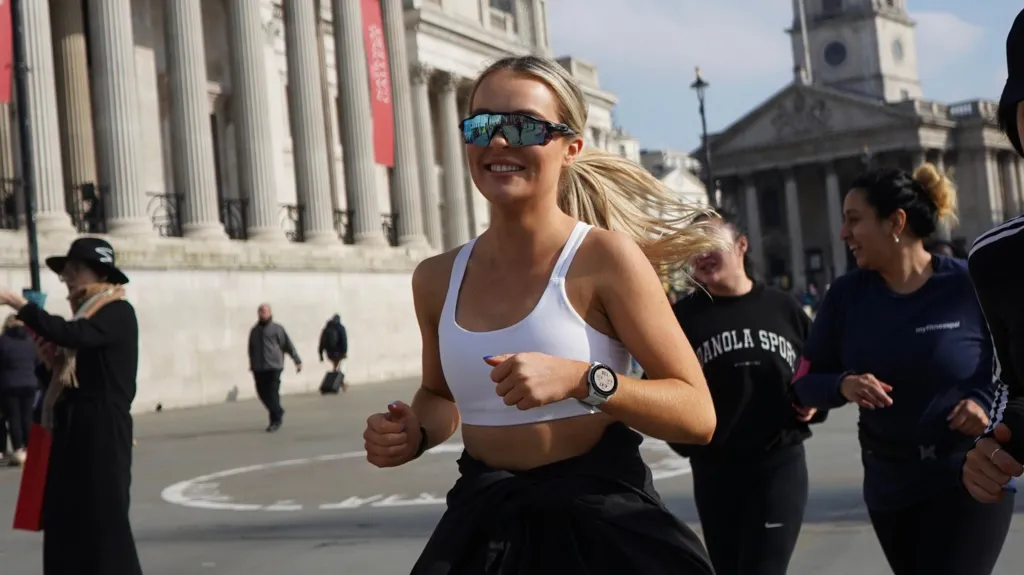 Maddie, who has long blond hair tied back, is on a run with other people. She wears a white top, smart watch and sunglasses - there are buildings in the background. 