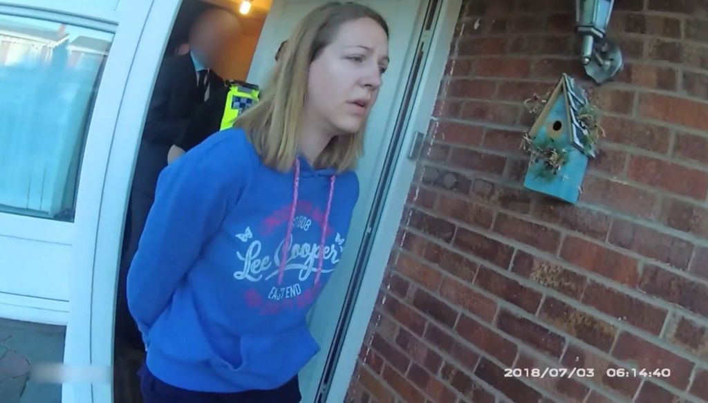 Lucy Letby shown on police body cam being arrested and taken from her home. She is wearing a blue Lee Cooper hoody and is walking past a brick wall with a bird box on it. The date read 2018/07/03 and the time is 06:14:40. She is followed by a police officer and a plain clothes detective who are blurred out.