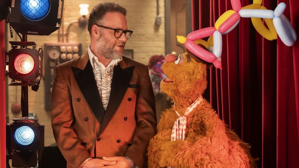 Seth Rogen wears a 70s style smoking jacket and ruffled shirt as he stands with his hands folded in front of him next to studio lights - he is talking to puppet Fozzie, who wears an elaborate balloon hat 