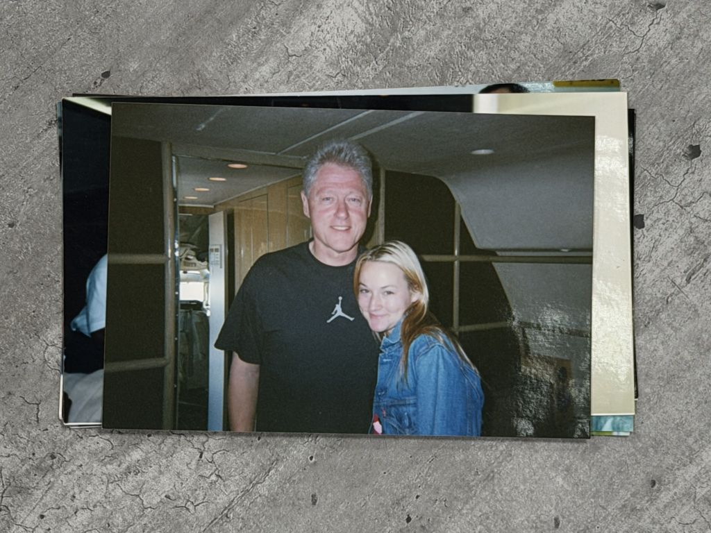 Bill Clinton, an older man with short grey hair, stands close to a young woman, Chauntae Davies. Clinton is in a black short-sleeve t-shirt that has the Air Jordan logo on it. Davies is wearing a light-washed jean jacket. They are inside a plane.