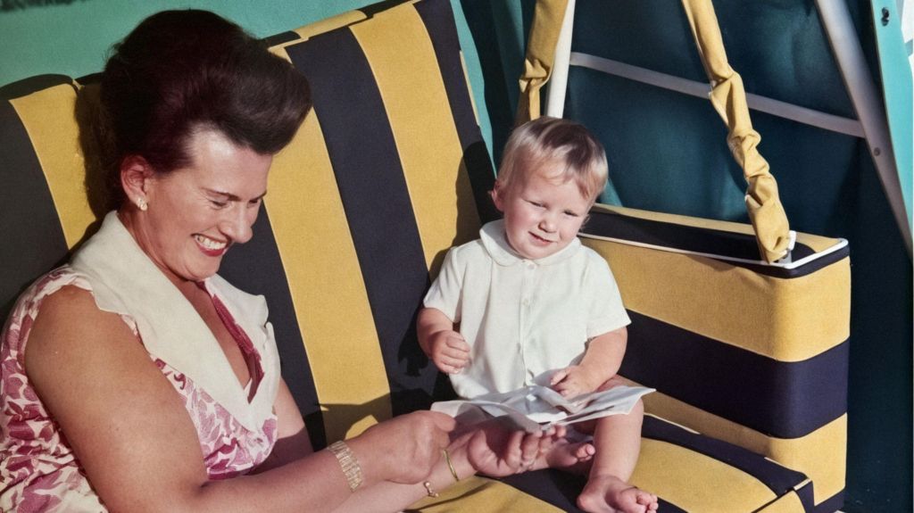 Muriel McKay sitting on a striped black and yellow garden swing with her grandson Mark as a young child. She is giggling and holding a book or piece of paper, which he is looking at. He wears a white collared t-shirt and has short blonde hair.