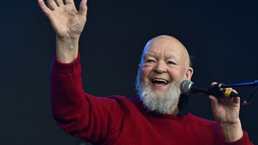 Sir Michael Eavis in a red jumper holding a microphone on stage.