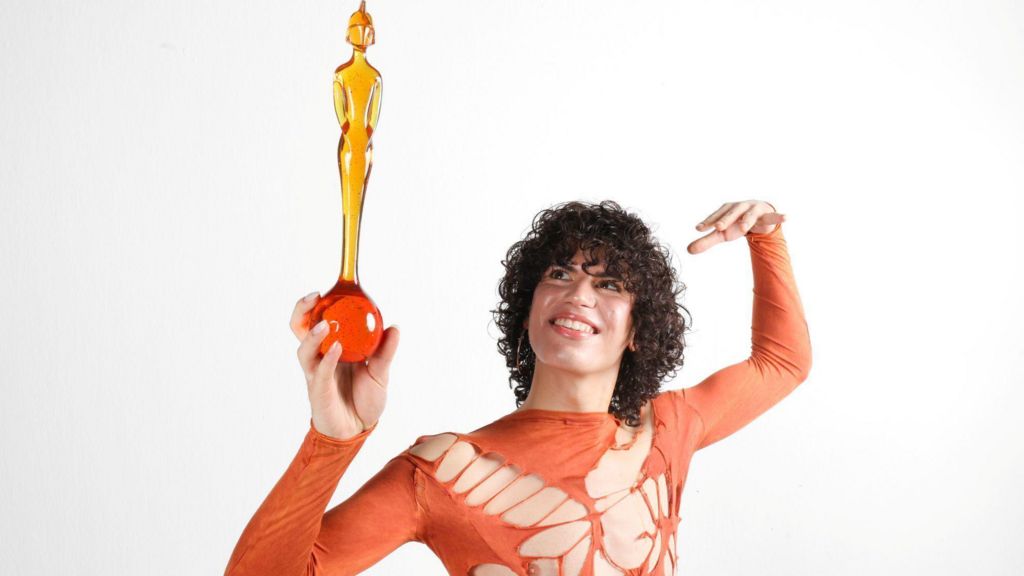 Jacob Alon is looking up at a Brit Award which they are holding in their hand. They wear a orange ripped top and have brown curly hair.