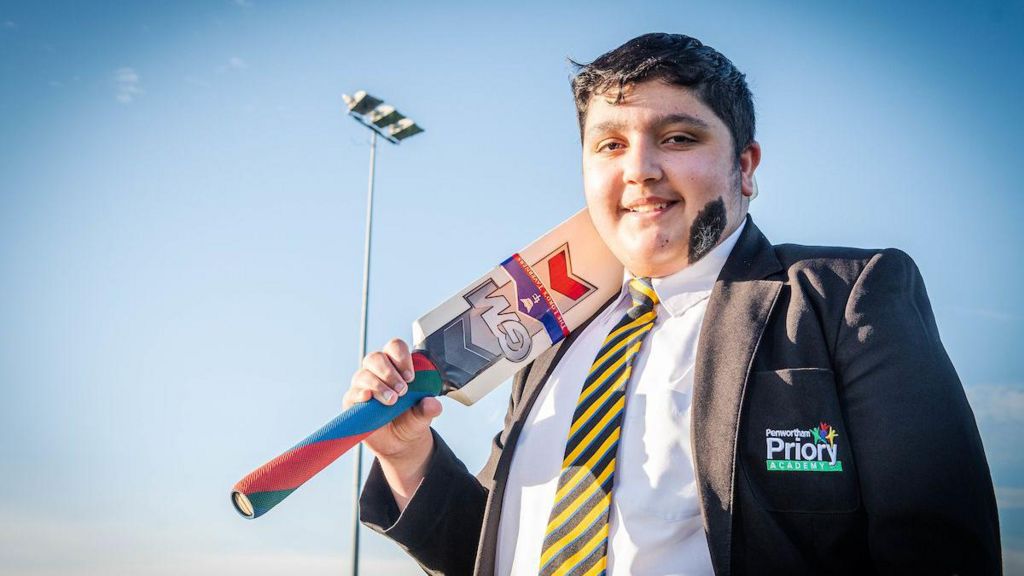 Umar Mahmood smiles at the camera and is wearing school uniform while holding a cricket bat.