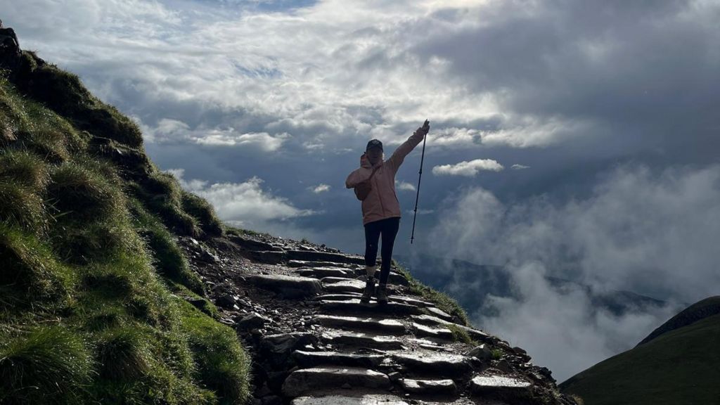 Sarah de Lagarde on a hike. She is standing along some stone steps on the side of a mountain, blue sky and clouds behind her and a grassy mountainside on the left.