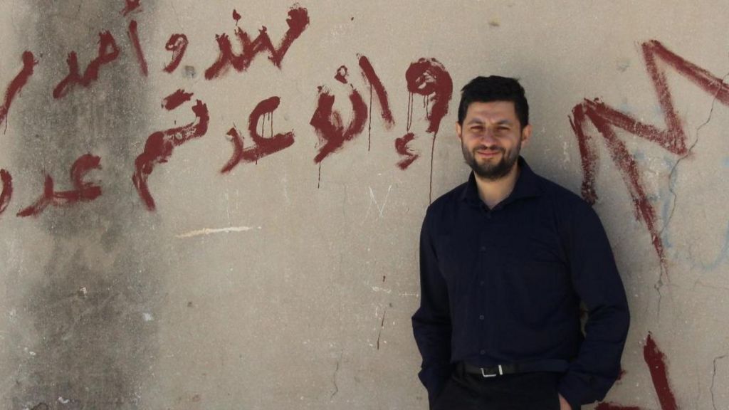Ammar Azzouz, a Middle Eastern man with a short, dark beard and short, dark hair, standing in front of a wall and smiling at the camera. The wall behind him has some writing in Arabic in red paint.