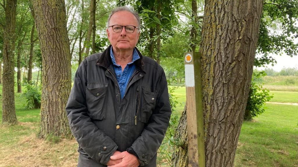 Henry Manisty, who has grey hair and glasses, standing among trees in Waterstock. On a wooden post behind him an orange arrow points towards a public footpath.