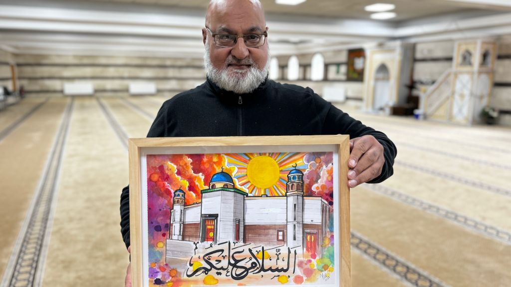 A man wearing black holds a hand painted picture of a mosque while standing in a worship room.