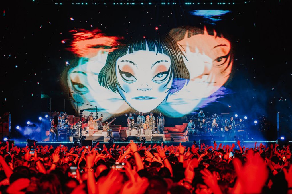A shot of Gorillaz stage show in Tottenham, shot from the crowd. Dozens of on-stage musicans are seen underneath a large animated image of a disembodied face