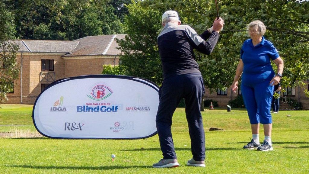 Guide Di Daniels, wearing a blue tracksuit, looks on as her husband Danny Daniels, in golfing gear, takes a swing with his club at the British Blind Masters tournament