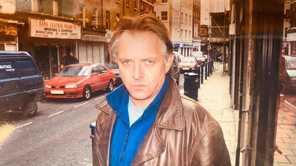 Rik Mayall, with mid-brown hair and grey streaks on the side which is swept back, wearing a brown leather jacket over a royal blue fleece and a light blue t-shirt. He is standing on a city street with cars going by and looking at the camera with a stern expression on his face.