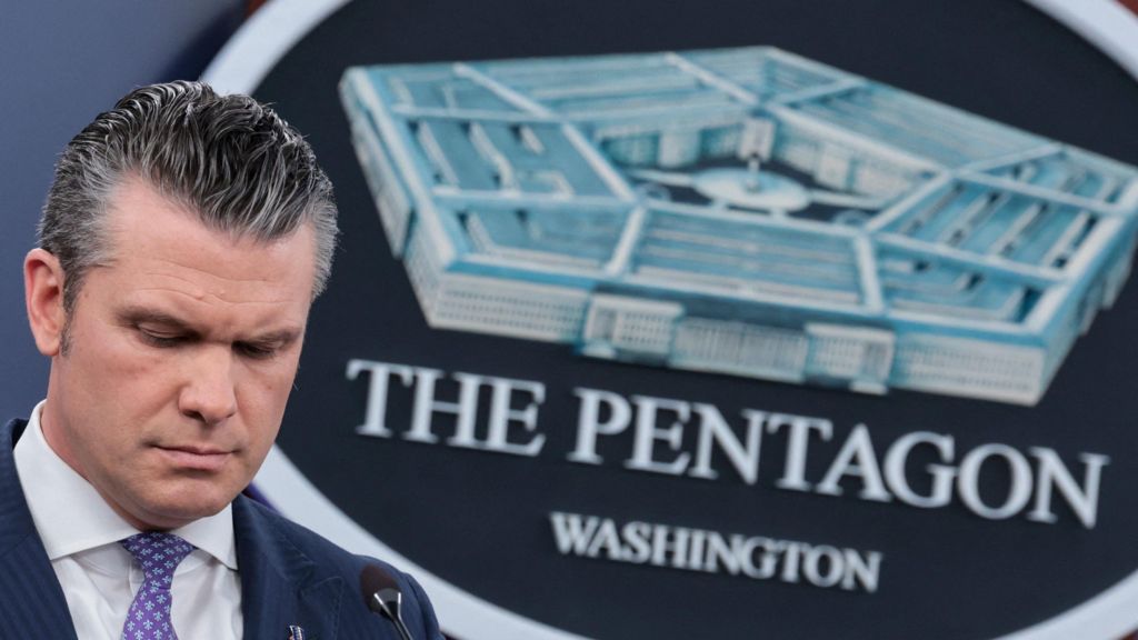 US Secretary of Defense Pete Hegseth, wearing a blue suit jacket, purple and blue tie, and a white dress shirt, stands beside a logo for the Pentagon as he looks downward.