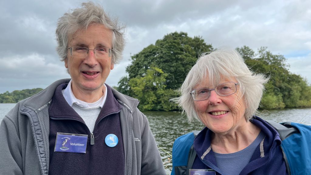 A man and woman smile at the camera. The man is wearing a grey jacket with a navy fleece underneath and a badge that says volunteer. The woman is wearing a blue jacket. Behind them is a body of water with a cluster of trees in the middle