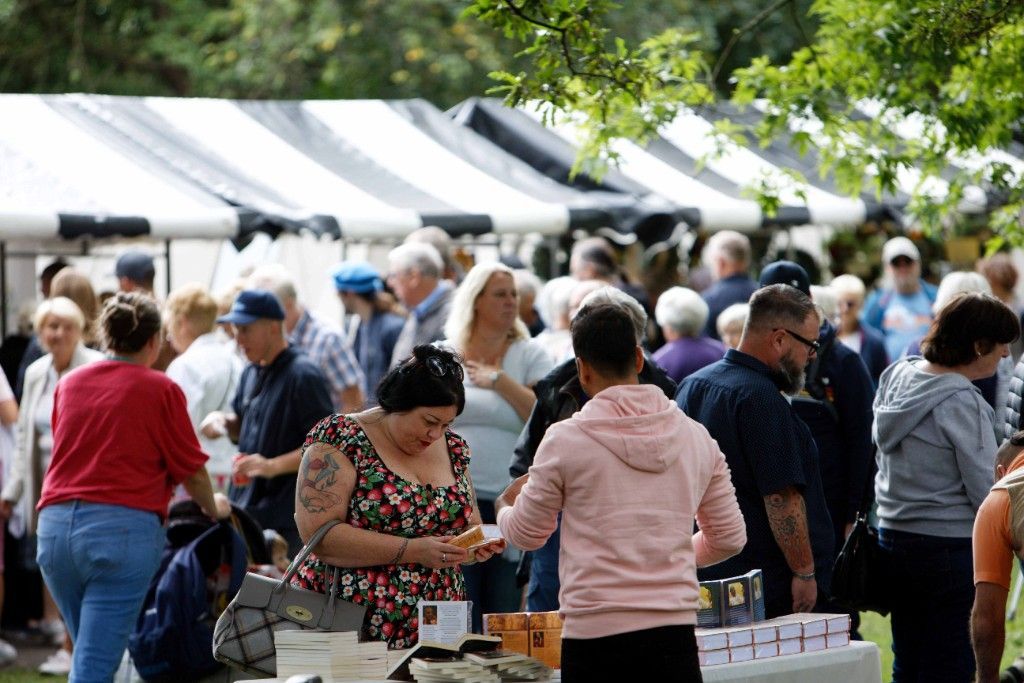 A woman in a flower-patterned dress is buying good from a market trader at an event where a crowd of people and tents can be seen in the background