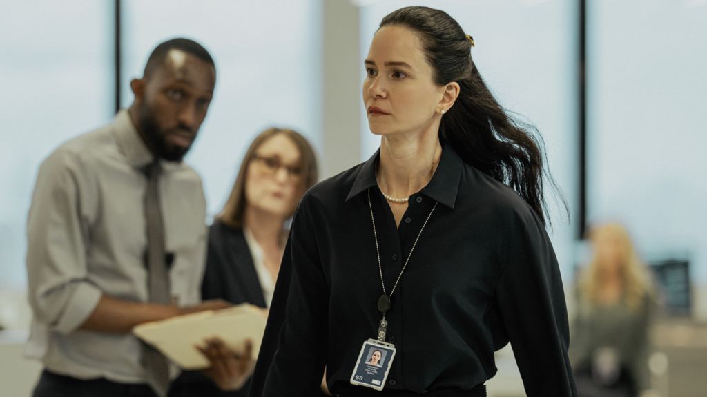 Katherine Waterston walks around an office wearing a black shirt and lanyard with people in the background staring at her