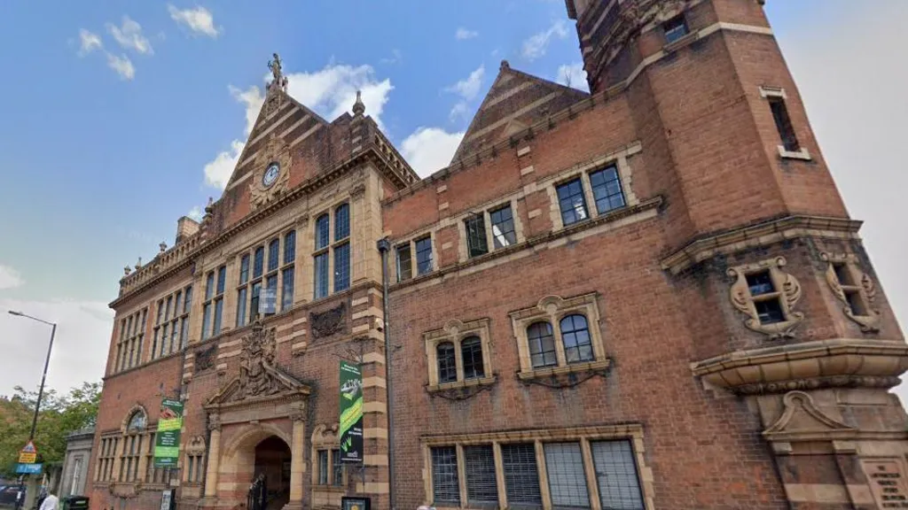 A large red brick building with small, black framed windows and people walking outside. There is a blue sky overhead with the odd white cloud.
