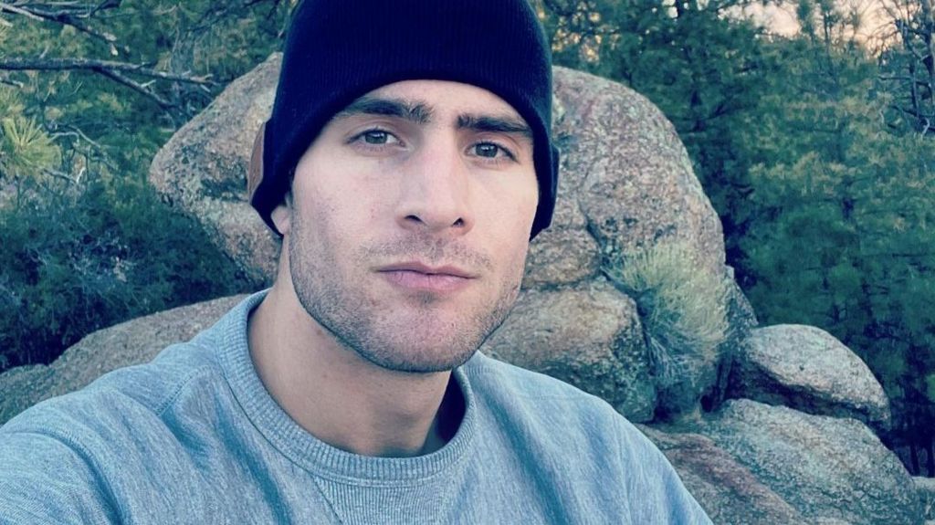 Keith Milkie, a young man wearing a grey sweatshirt and a dark beanie, pictured on a rocky outcrop with a forest behind him