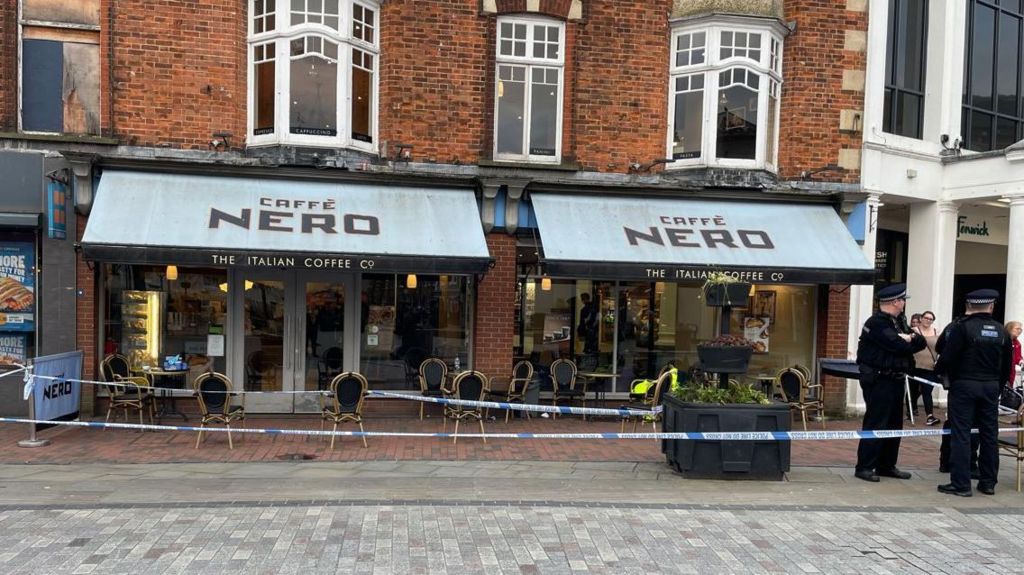 The front of a brick built Cafe Nero cafe featuring blue awnings and police tape extending from the cafe across the nearby pavement